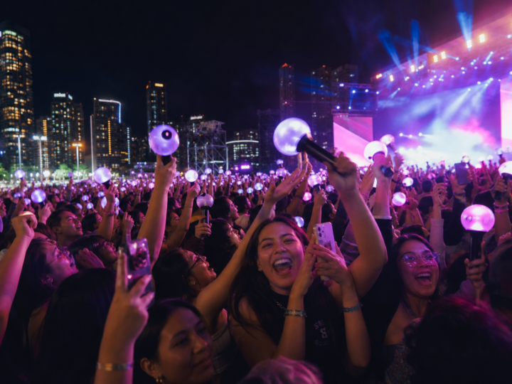 Latin American pop fans at a night concert holding light sticks and phones in a colorful crowd.